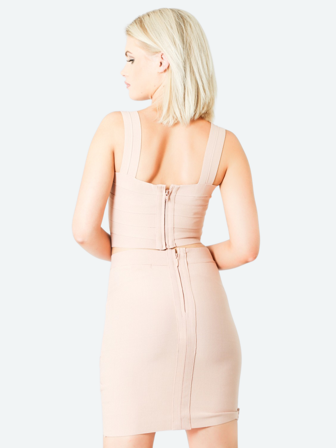 Model wears a bandage crop top with  a criss cross design, stretch fit and zip back fastening in nude colour. The model wears a matching bandage skirt to complete the look. The model has blonde hair and stands with her back to the camera. he back of the outfit can be seen, including the zip back fastening of the top and skirt. 