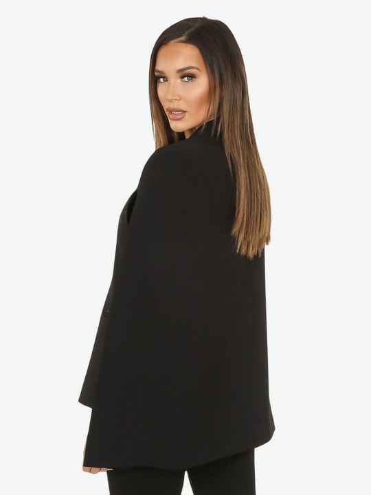 Model wears black cape blazer with side split and black satin lapel. The blazer has front button fastening.  Model stands with her back to the camera, the cape structure of the cape can be seen. Model wears glamorous makeup.