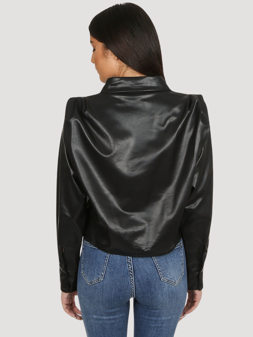 Model wears black faux leather shirt with pleated shoulders and long sleeve. The pu cropped front button shirt and collard neck. Model stands with her back to the camera. The back of the cropped pu leather shirt, the pleated puff shoulders are visible.