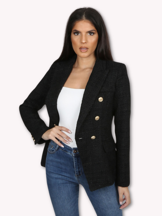 Model wears a black metallic yarn knitted double breasted blazer. The double breasted blazer has gold buttons at the front of the blazer and at the cuffs. Model stands with one hand on her hip and the other by her side, She stares directly into the camera with her mouth slightly open. 