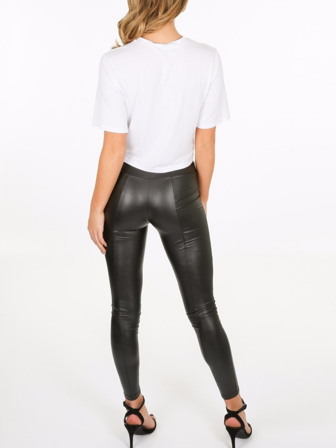 Whole body shot. Model wears a wet look, leather looking leggings with panelling. This faux leather leggings has a bodycon slim fit.  Model stands with her back to the camera with one hand in front and the other behind her back. The back of the panelled wet look leggings can be seen. 