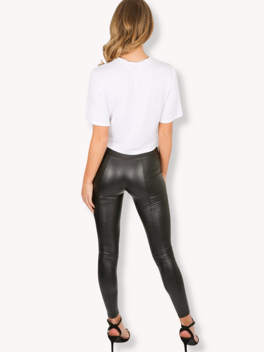 Whole body shot. Model wears a wet look, leather looking leggings with panelling. This faux leather leggings has a bodycon slim fit.  Model stands with her back to the camera, the back of her panelled leggings can be seen.