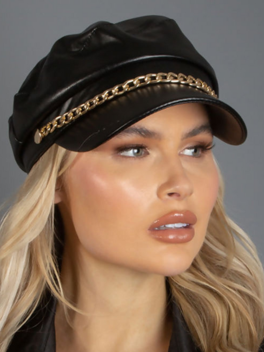 Model wears a faux leather baker boy hat with gold chain detailing at the front. Model looks off into the distance, the front of the cap can be seen. 