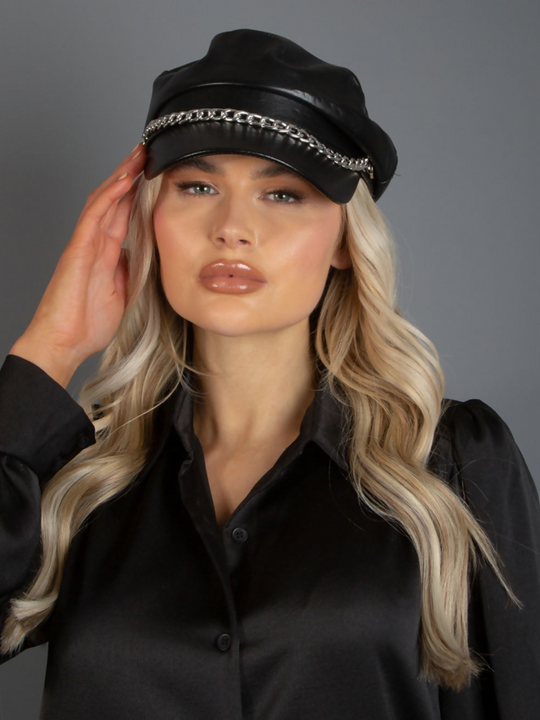 Model wears PU leather black baker boy hat with a silver chain detailing at the front. Model faces the camera and places one hand to the side of the hat. 