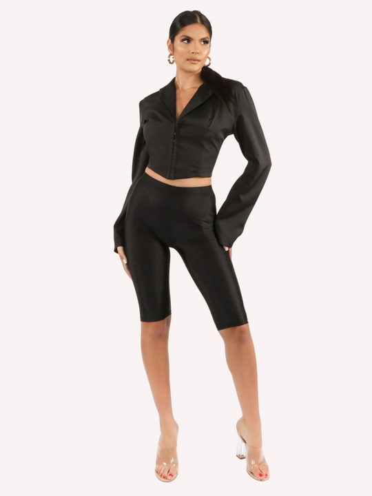 Full length shot. Model wears a black cropped shirt with a lapel, hook eye front closure and long sleeves that are wider at the cuffs.  Model faces forward with one hand slightly in front of the other and her hands by her sides. Model pairs the cropped shirt with black cycling shorts.