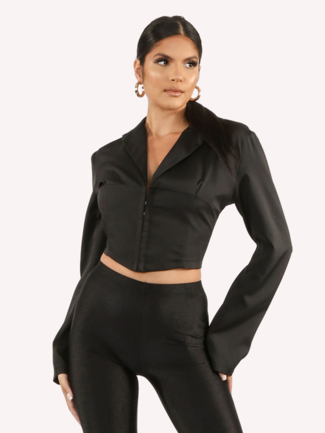 Model wears a black cropped shirt with a lapel, hook eye front closure and long sleeves that are wider at the cuffs.  Model faces forward, standing with both hands on her hips and stares off into the distance. 