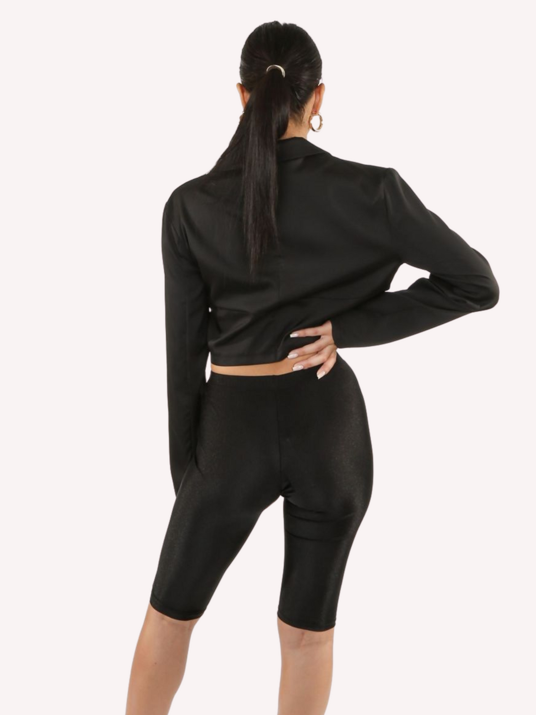 Model wears a black cropped shirt with a lapel, hook eye front closure and long sleeves that are wider at the cuffs.  The model stands with her back to the camera, the back of the shirt can be seen. 