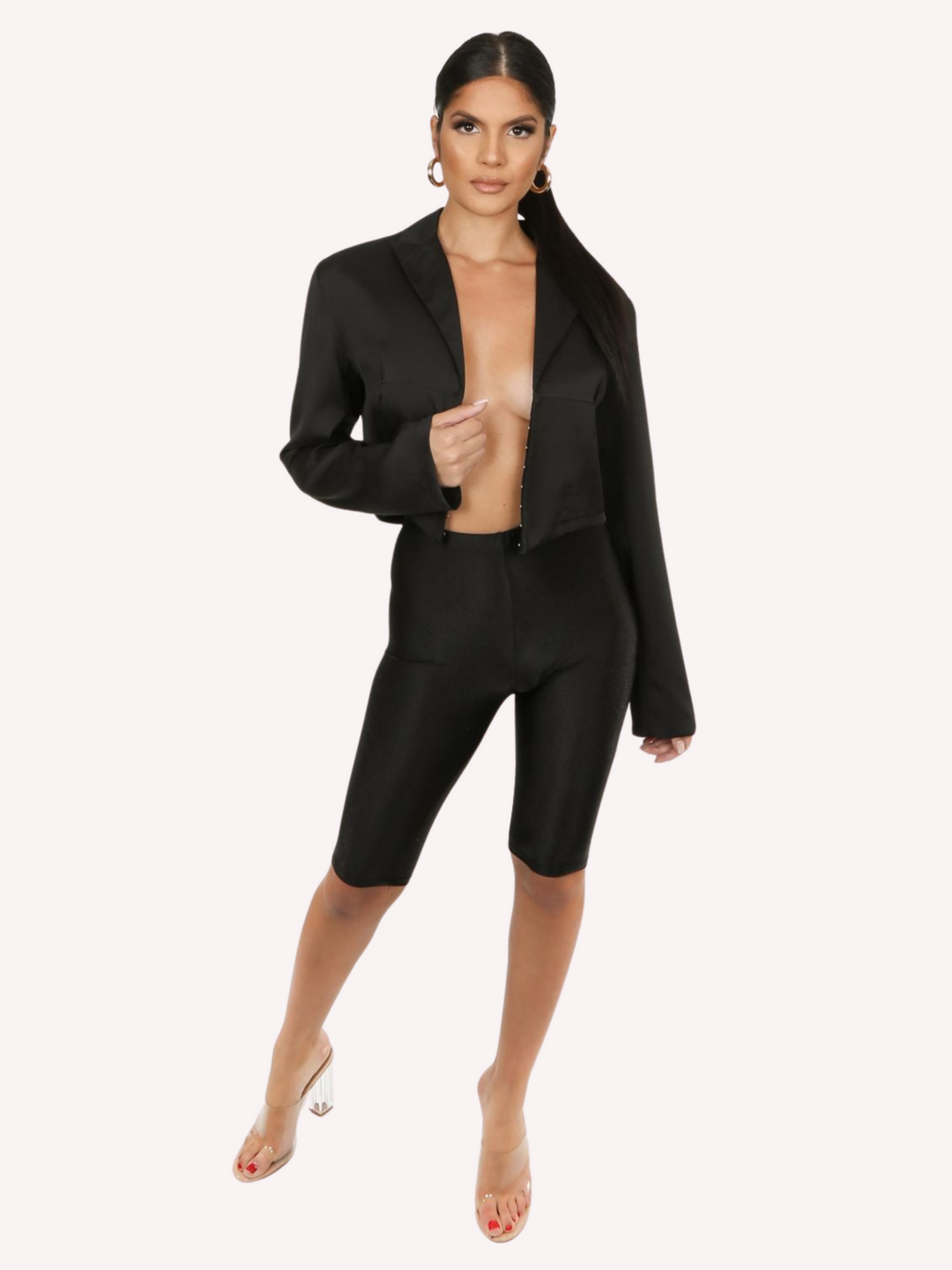 Full length shot. Model wears a black cropped shirt with a lapel, hook eye front closure and long sleeves that are wider at the cuffs. Model stands with the shirt open and faces the camera.