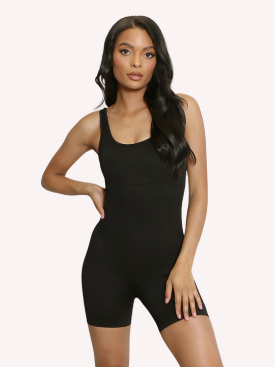 Model wears a black ribbed unitard jumpsuit with a scoop neck, waist control and open back. Model stands with one hand on her hip and one hand on her thigh.