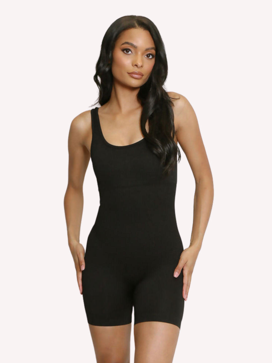 Model wears a black ribbed unitard jumpsuit with a scoop neck, waist control and open back. Model stands with both hands on her hips.