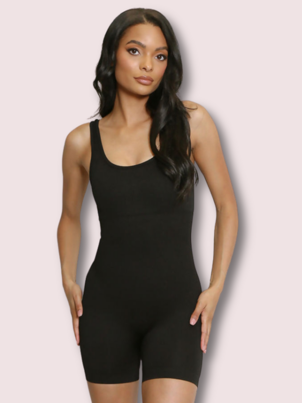 Model wears a black ribbed unitard jumpsuit with a scoop neck, waist control and open back. Model stands with both hands on her hips.