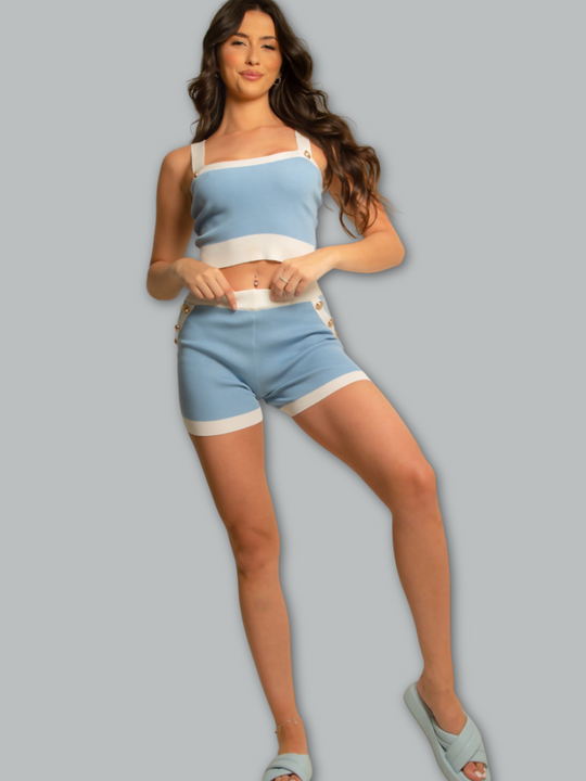 Full length body shot. Model wears a baby blue and white fine knit shorts set, with matching blue and white crop top and and shorts and gold military buttons. Model stands with one leg to the side and her hands on the short's waist band.