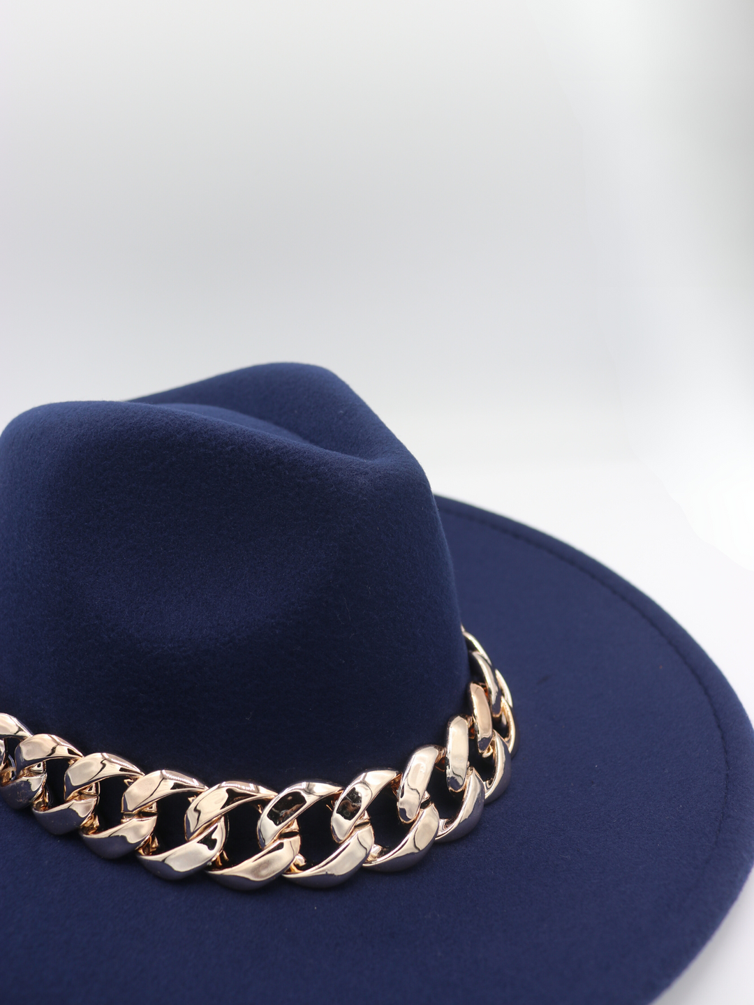 The side and front of the hat can be seen with an empahsis on the chunky gold chain. The hat is of a luxe navy felt fedora hat with chunky gold chain design at the centre of the hat.