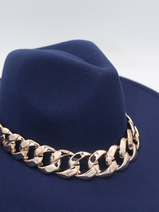 Close up of  the top of the navy felt fedora hat can be seen including the indentation and the gold chunky linked chain detailing.