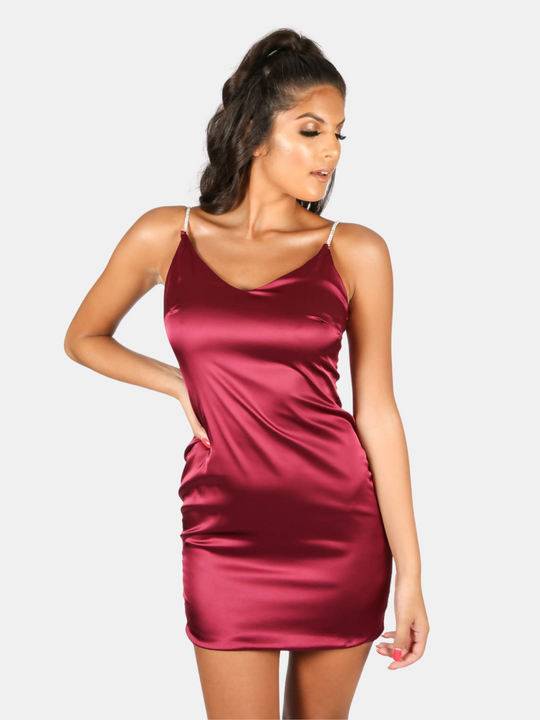 Model wears a red wine coloured satin mini dress with diamante straps and concealed zip back fastening. Model stands with one hand on her hip and the other by her side. The front of the dress can be seen. 