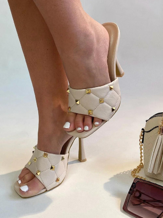 Cream quilted faux leather mule heels with gold studded heels. Model waers cream studded mule heels, she lifts obe leg above the ground. A hand bag and sunglasses are on the ground. 