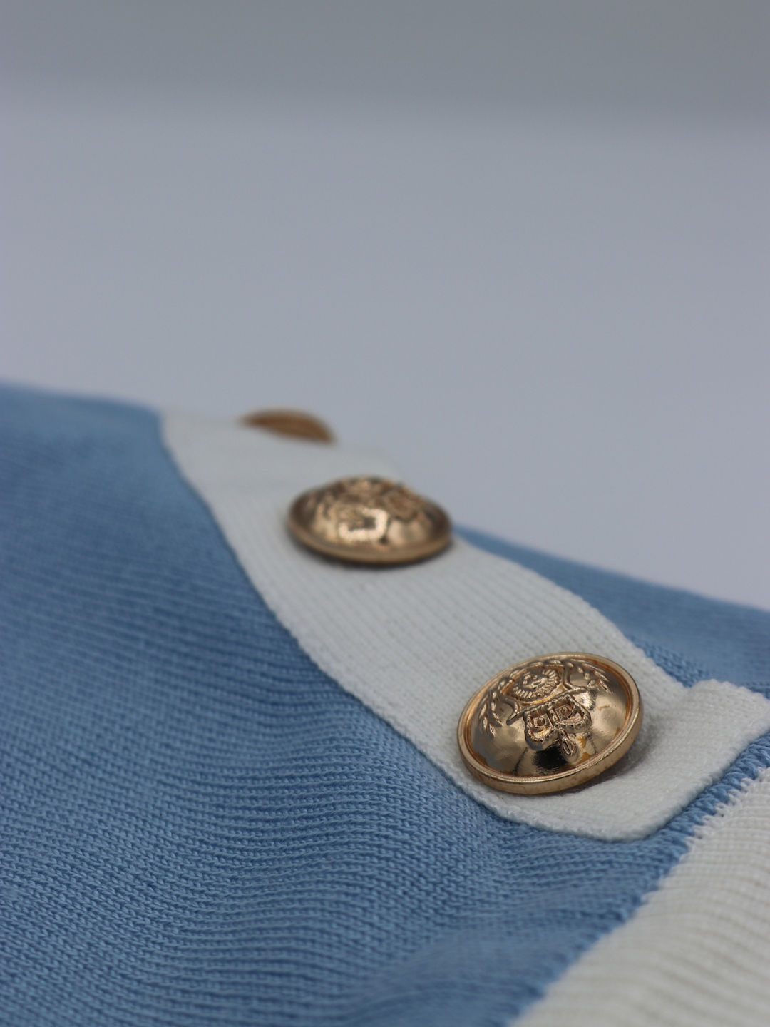 Close up of fine knit baby blue co ord set. A close up of the gold  military button detailing can be seen. The gold buttons sit on the white knit design.