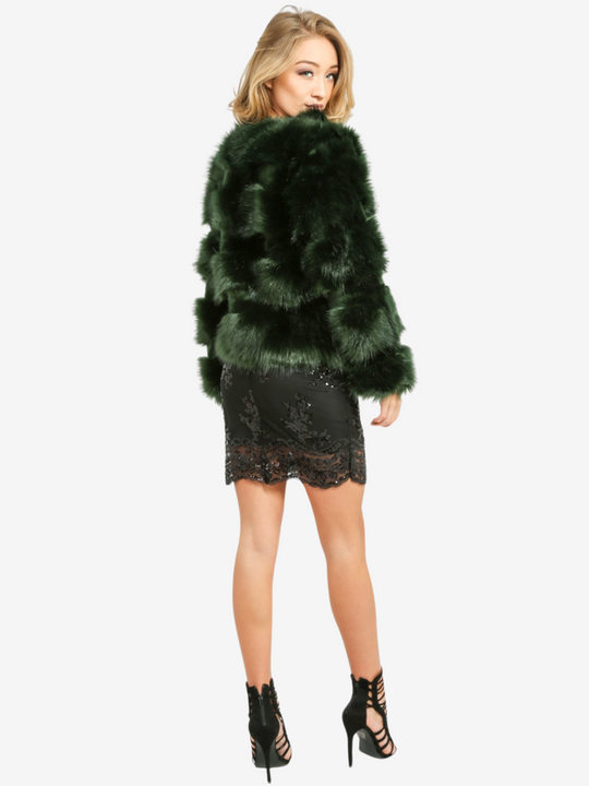 Full length shot. Model wears a soft faux fur long sleeve green jacket. The jacket has a front fastening and  satin like inner lining. Model stands with her back to the camera,  her sequin black dress, black high heels and the back of the faux leather jacket can be seen. Model turns her head sp that she is looking at the camera.