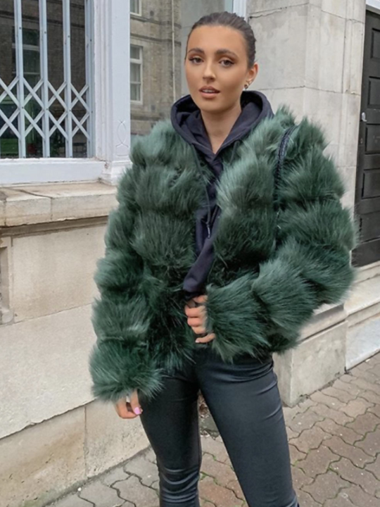 Model stands in the street. She wears a soft faux fur long sleeve green jacket. The jacket has a front fastening and  satin like inner lining. Model wears the jacket open,  she wears a blue shoody and black skinny fit jeggings. 