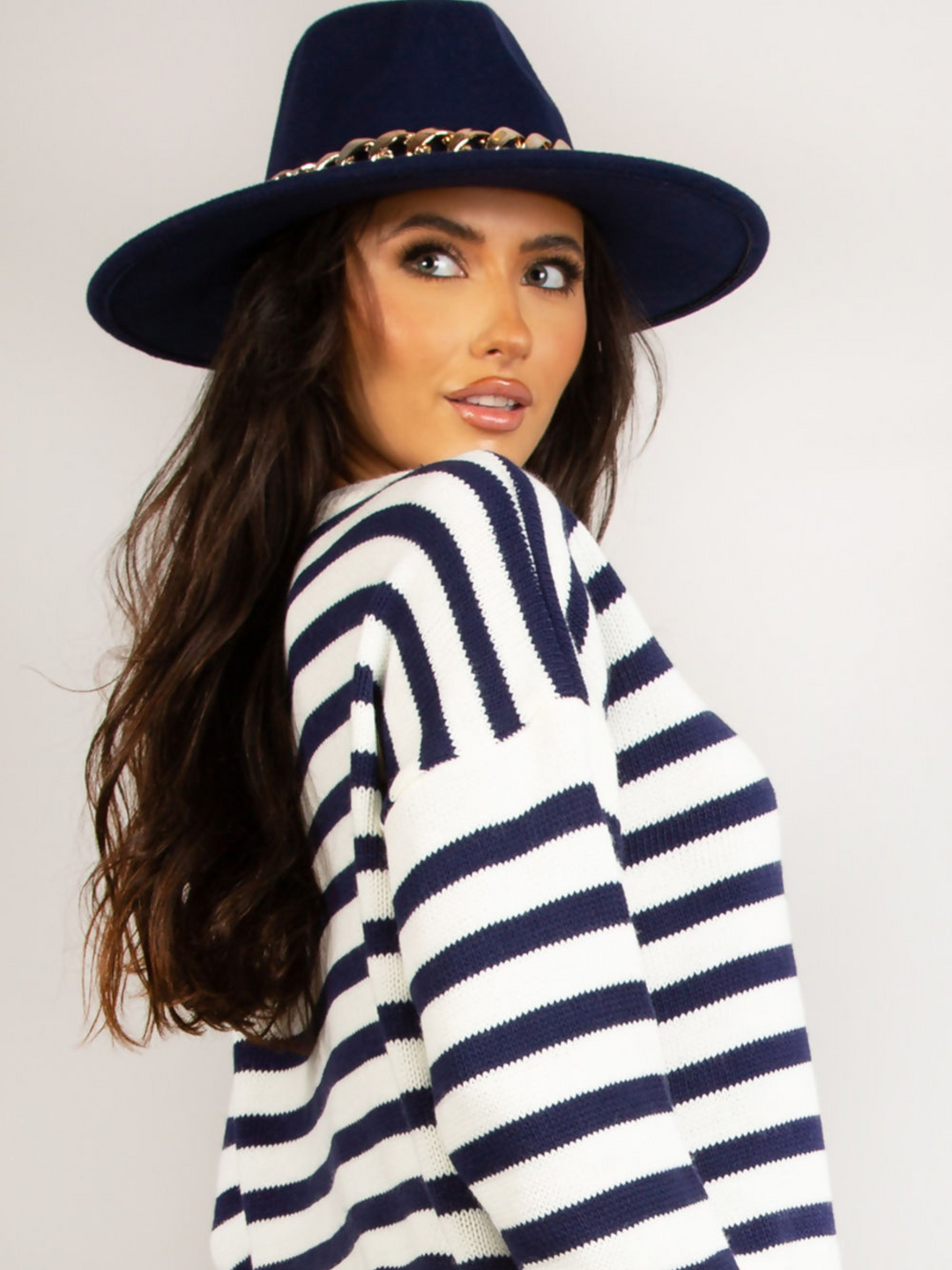 Model wears a luxe navy felt fedora hat with chunky gold chain design at the centre of the hat. Model stands to the side and looks off into the distance, the front of the hat can be seen. 