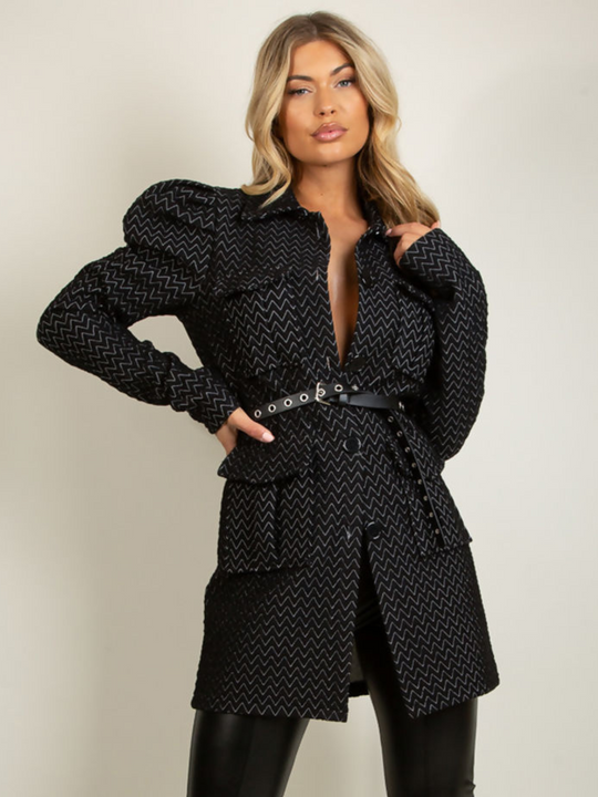 Model wears a black longline blazer with soft quilted patterns with a soft textured finish. The shoulders have a puffed finish with long sleeves and front button fastening. The model wears a belt that  is fed between the belt loop. Model places one hand on her hip and one hand on her shoulder. 