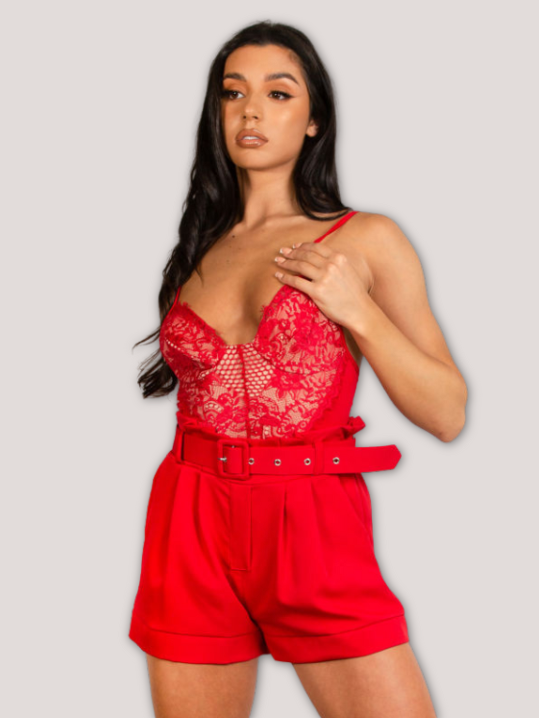 Model wears a red bodysuit with lace panel detailing. Model stands with one hand behind her back anf the other holding the tops spaghetti straps. 