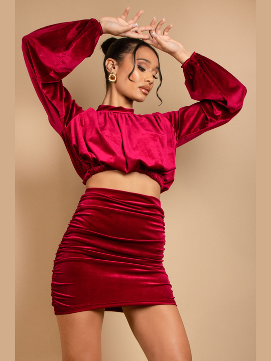 Model wears a long sleeeved red velvet crop top with a high neck. She also wears a matching  ruched mini skirt. The model faces forward and places her hands above her head and looks down. 