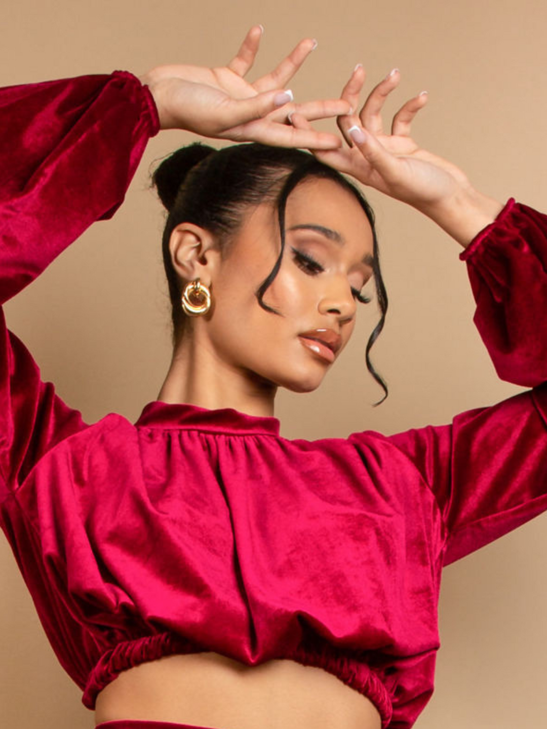 Closeup of the cropped top. Model wears a long sleeeved red velvet crop top with a high neck. She places her hands above her head and looks down.