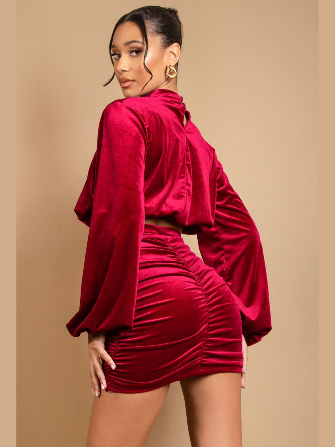 Model wears a long sleeeved red velvet crop top with a high neck. She also wears a matching  ruched mini skirt. Model stands with her back to the camera. The back of the outfit can be see, de,onstrating the button fastening of the crop top and ruched bum design of the stretchy skirt.