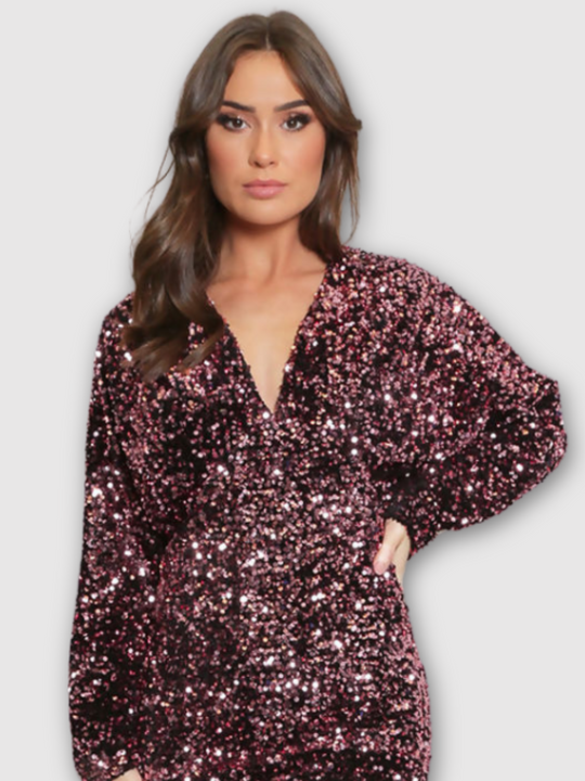 Model wears deluxe rose sequin mini dress with long sleeves. The neck has a v neck and concealed back zip fastening. Model faces forward and places one hand on her hip. The sequined dress shows its shimmer. 