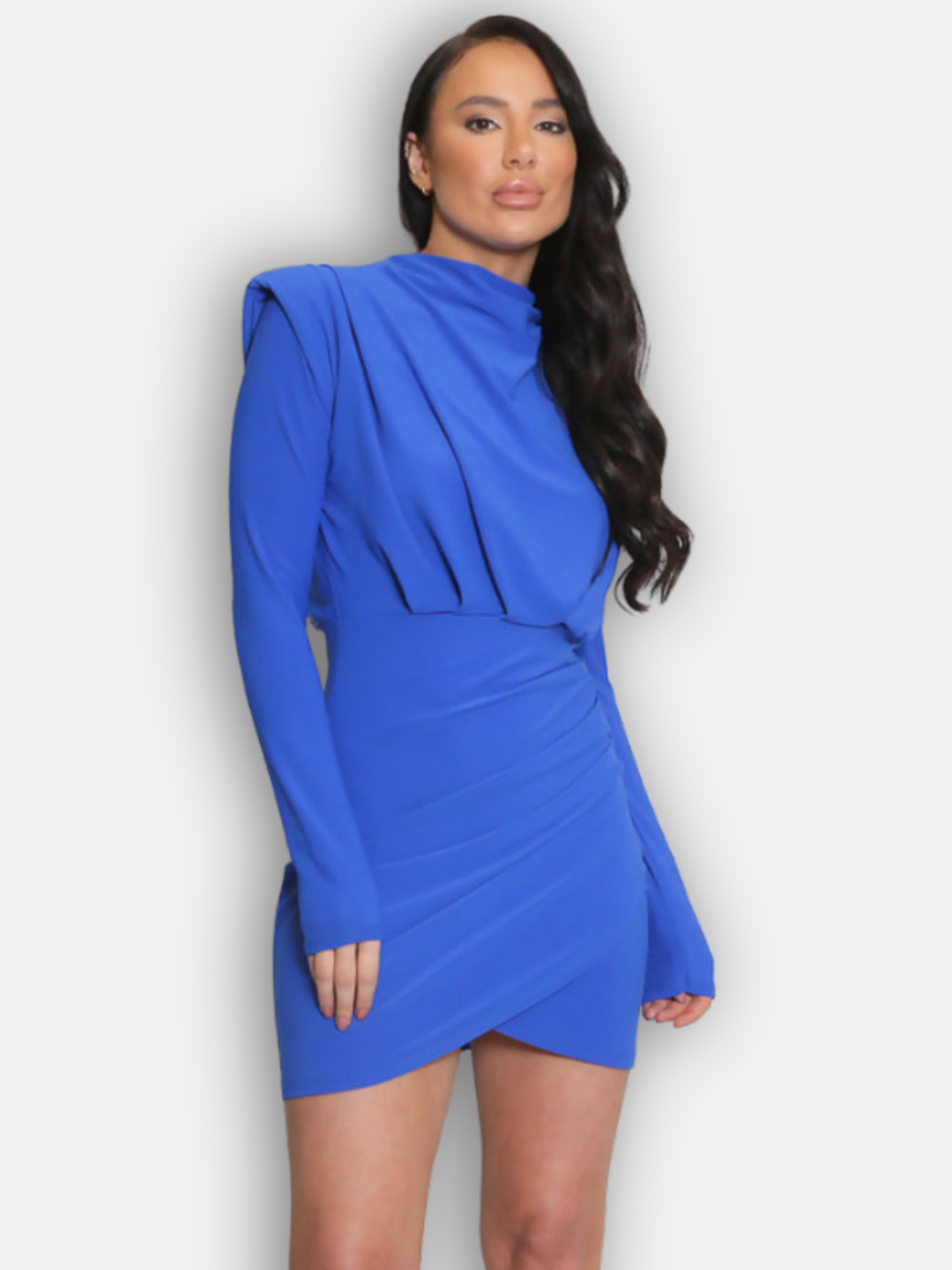 Model wears blue mini dress with long sleeves and shoulder pads. Model stands facing forwards with boths hands by her sides. Model has her wavy hair styled to one side. 