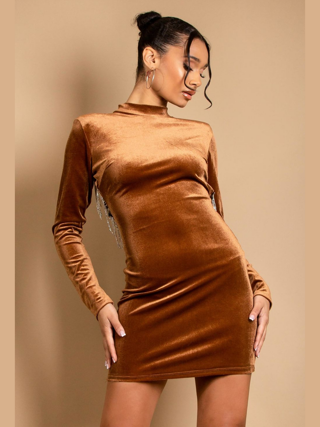 Model wears a velvet mini dress in a tanned colour. The mini dress has a bodycon fit and velvet elastic, with a mock neck, long sleeves and a cut out back with diamante trim. Model faces forward with her hands by her sides. The diamante tassles situated at the back can ne seen hanging down. Model looks down. 