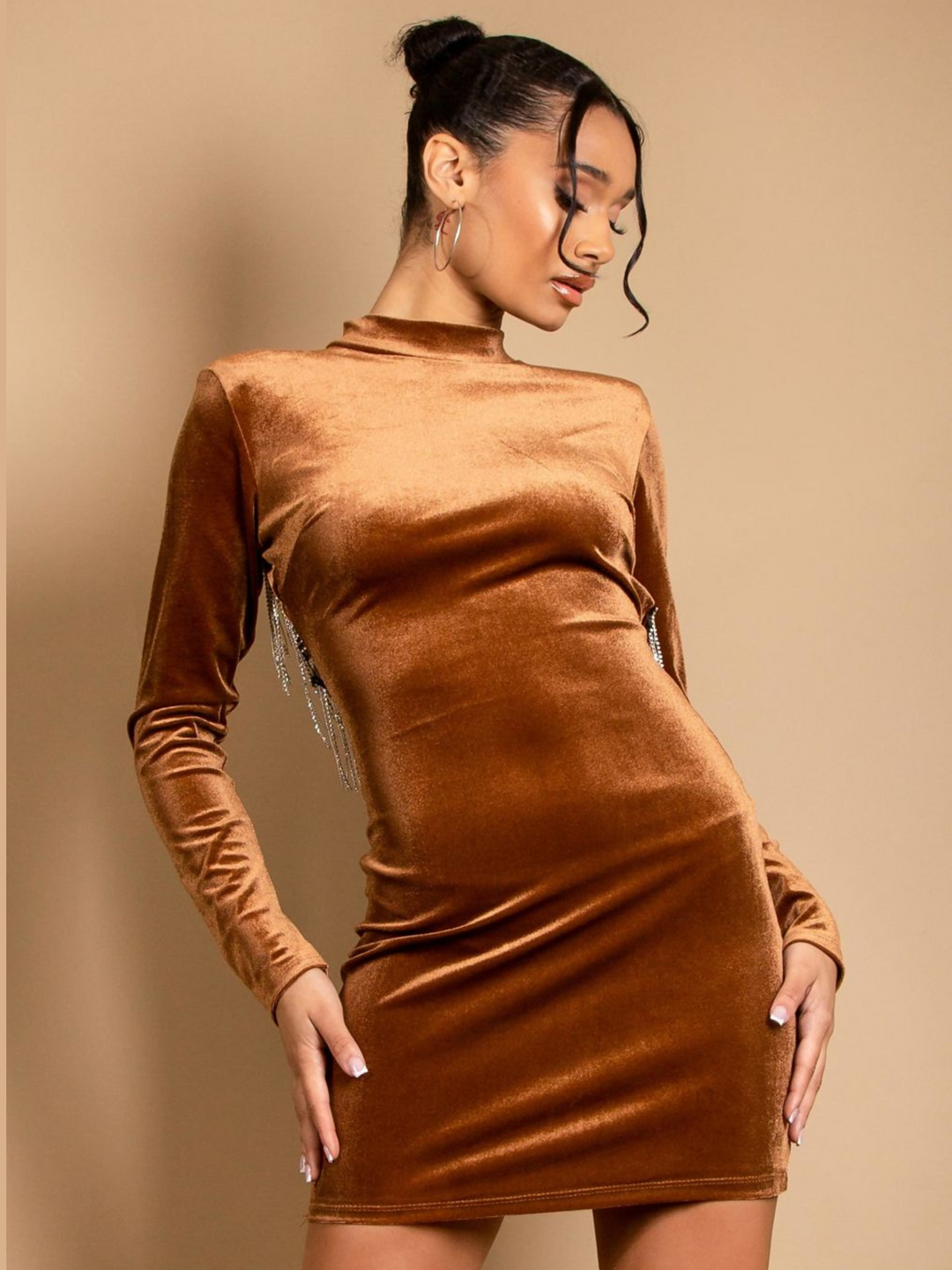 Model wears a velvet mini dress in a tanned colour. The mini dress has a bodycon fit and velvet elastic, with a mock neck, long sleeves and a cut out back with diamante trim.  Model faces fprward and looks away from the camera.