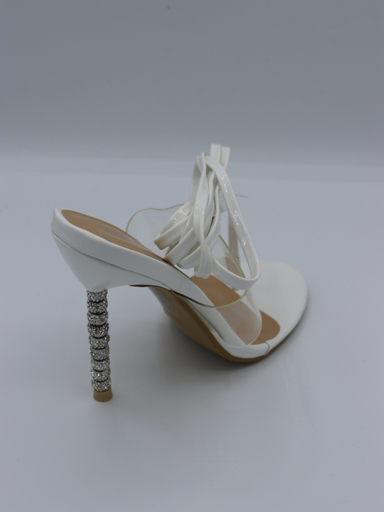 White strappy mules shoes with diamante stiletto heel. The perspex strap that goes across the shoe is visible, as is the white straps that can be used to go up the legs or around the ankles. 