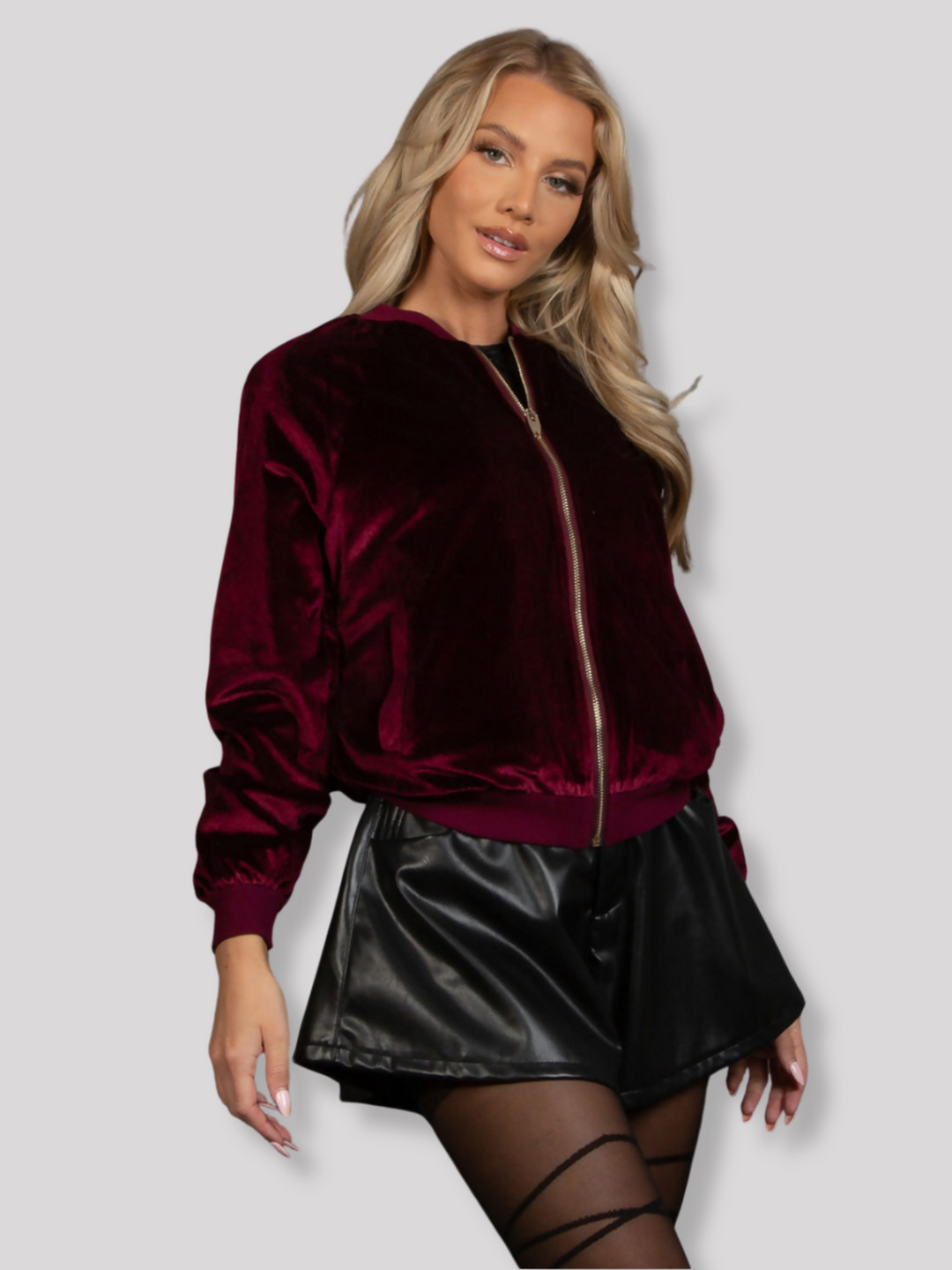 Model wears a red wine coloured velvet bomber jacket, that has long sleeves,  a front zip fastening and rounded neck.  Model stands at an angle and looks into the camera, her hands are by her side and the jacket is zipped up. 