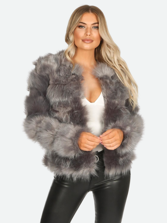 Model wears graphite grey faux fur jacket with long sleeves, rounded neck and front closure fastening. Model faces the camera with both hands clasped. She places one leg slightly to the side. 