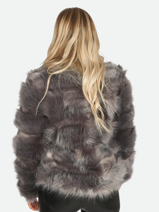 Close up. Model wears granite grey faux fur jacket with long sleeves, rounded neck and front closure fastening.  Model stands with her back to the camera, the back of the faux fur can be seen. 