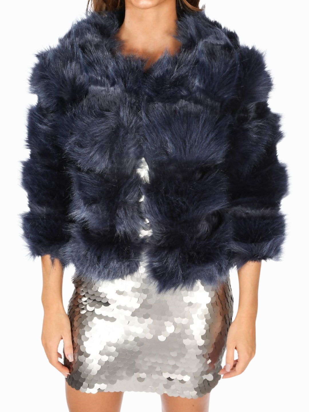 Mid length shot. Model wears a soft faux fur navy jacket with cropped sleeves and front clasp fastening at the top.  She wears a silver sequin mini dress with black high heels. The jacket has been fastened at the front. 