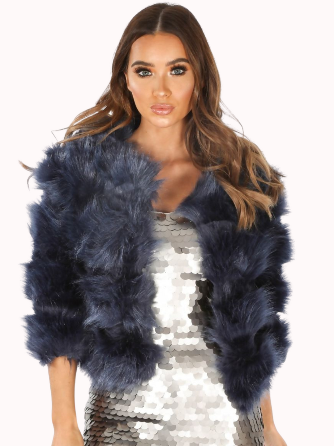 Mid length shot. Model wears a soft faux fur navy jacket with cropped sleeves and front clasp fastening at the top. Model wears  a silver sequin dress underneath the dress and wears the faux fur jacket open. The model faces the camera with both of her hands by her sides. 