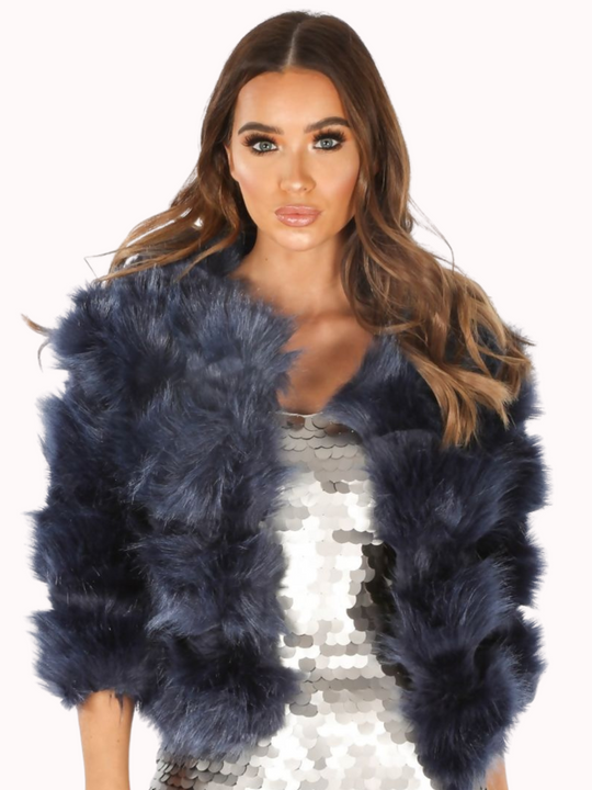 Model wears a soft faux fur navy jacket with cropped sleeves and front clasp fastening at the top. Model wears  a silver sequin dress underneath the dress and wears the faux fur jacket open. The model faces the camera with both of her hands by her sides.  Model has brown hair and blue eyes.