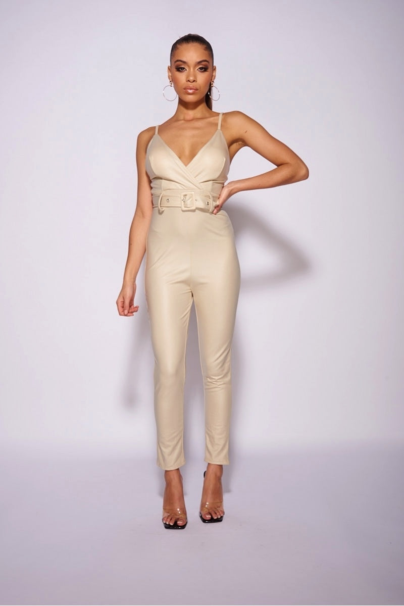 Model wears faux leather beige jumpsuit with high waisted belt . The jumpsuit has a skinny fit  style and a plunging neck line. Model stands with one hand on her hips.