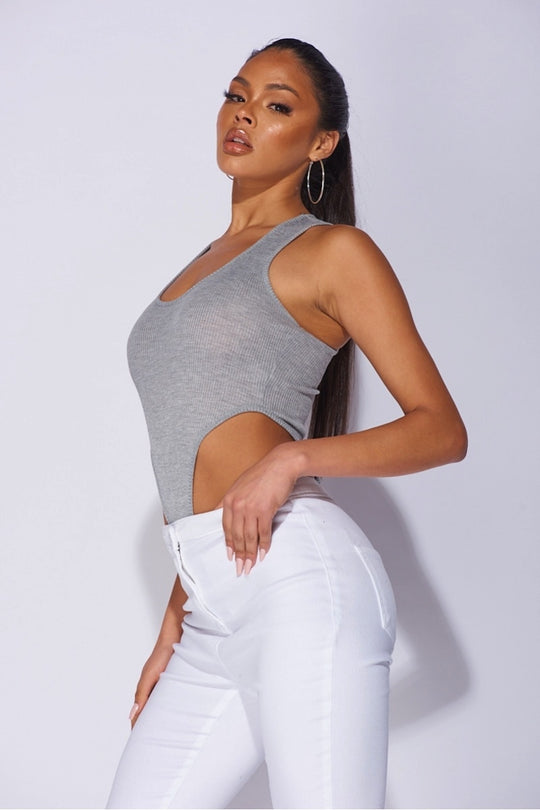 Model wears a high leg grey ribbed bodysuit with plunging neckline. Model wears white jeggings  Model stands to the side and has one hand on  her hips.