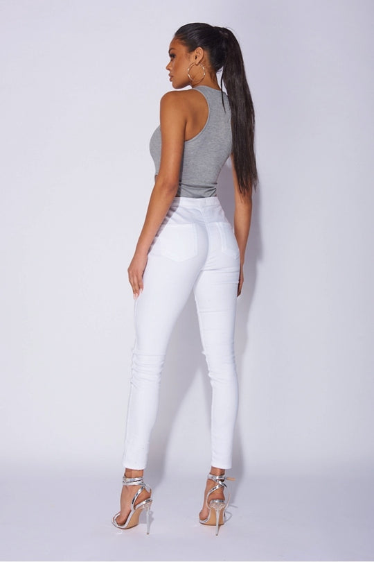 Model wears a high leg grey ribbed bodysuit with plunging neckline. Model wears white jeggings. Model wears silver stilletto heeled shoes and her hair is in a long pony tail. Model has her back to the camera and the racer back detailing of the bodysuit is visible.