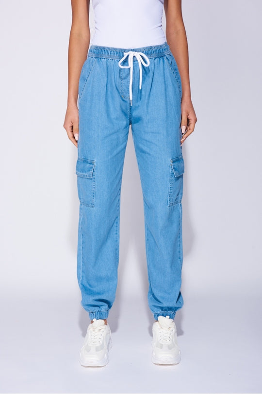 Model wears a loose fit light blue patch pocket cuffed denim joggers. Model wears a white bodysuit and white trainers.  Photograph shows a closeup of tghe trousers with the model with her hands behind her side. 